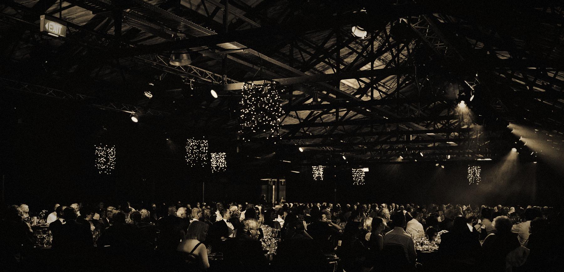 A large audience beneath a lit truss ceiling